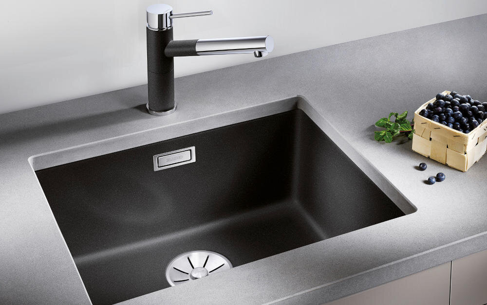 Czarny zlewozmywak jednokomorowy wbudowany w szary blat, z nowoczesną baterią. Obok znajduje się koszyk z ciemnymi owocami.