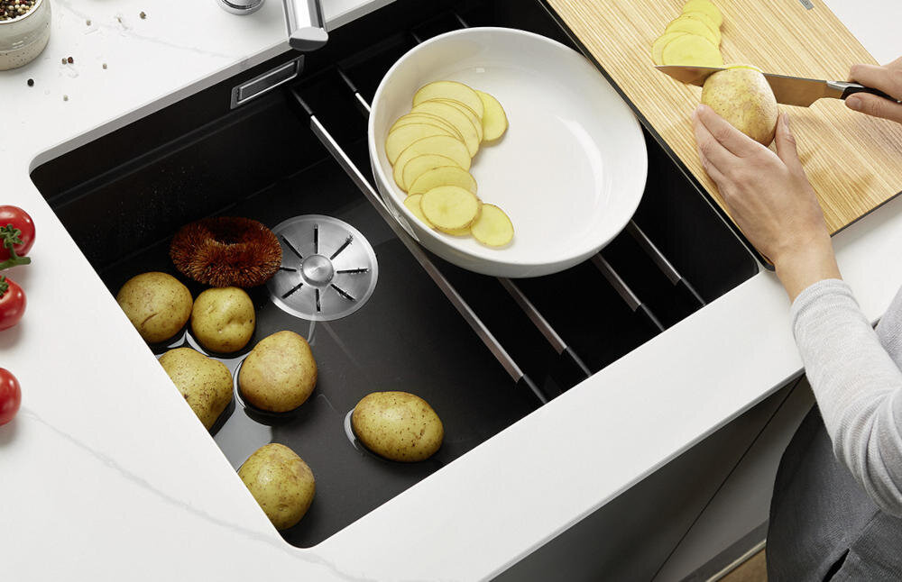 Na obrazie widoczny jest czarny zlewozmywak jednokomorowy z kilkoma ziemniakami w wodzie oraz osoba krojąca ziemniaka na desce do krojenia. Obok zlewozmywaka znajdują się pokrojone plasterki ziemniaka na białym talerzu.