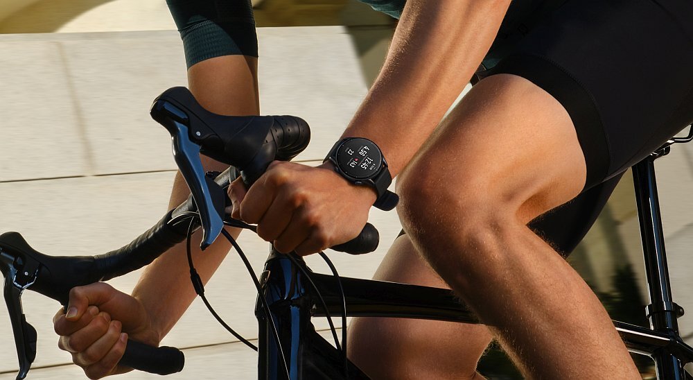 Osoba jedzie na rowerze, nosząc smartwatch na nadgarstku. Na ekranie zegarka widoczne są różne wskaźniki i dane.