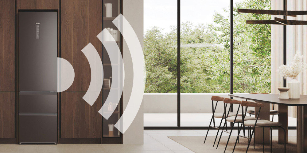 Wnętrze nowoczesnej kuchni z ciemną lodówką w zabudowie, obok której znajduje się stół z krzesłami. Na lodówce widoczne jest logo \'Haier\'.