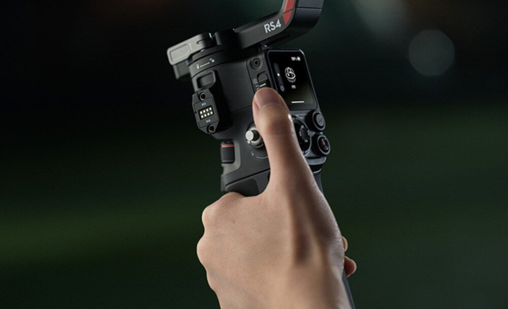 Osoba trzyma gimbal z widocznym napisem \'RS 4\' na ramieniu urządzenia. Na panelu sterowania widoczny jest ekran z ikoną oraz kilka przycisków.