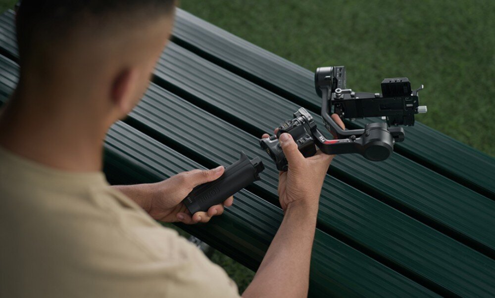 Osoba trzyma gimbal DJI RS 4, siedząc na ławce z zielonymi, metalowymi listwami. W tle widoczna jest trawa.
