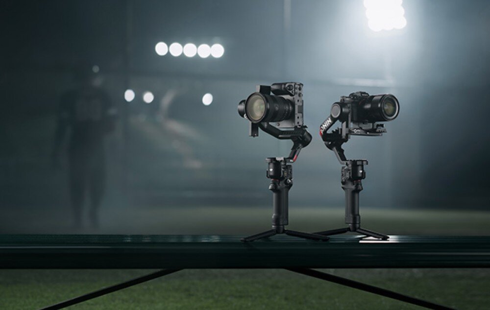 Na zdjęciu znajdują się dwa gimbale z zamontowanymi aparatami, ustawione na ławce w oświetlonym otoczeniu. W tle widoczna jest rozmyta postać oraz jasne światła.
