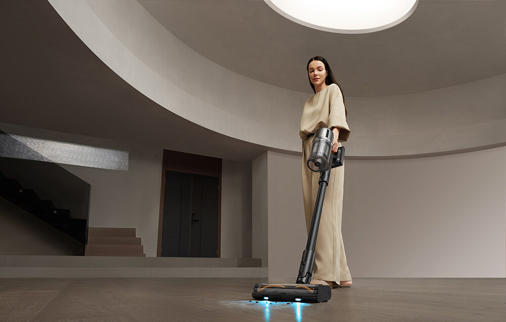 Osoba trzyma odkurzacz w nowoczesnym wnętrzu z dużym okrągłym otworem w suficie. Podłoga jest gładka i ciemna.