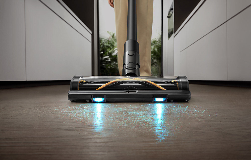 Odkurzacz z podświetleniem LED na końcówce ssącej, zbierający drobinki z podłogi. W tle widoczne są rośliny i fragmenty mebli.