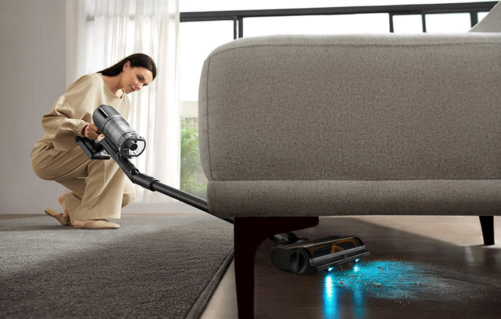 Odkurzacz z długą rurą ssącą czyszczący podłogę pod kanapą. Na podłodze widoczne są drobinki kurzu i światło emitowane przez odkurzacz.