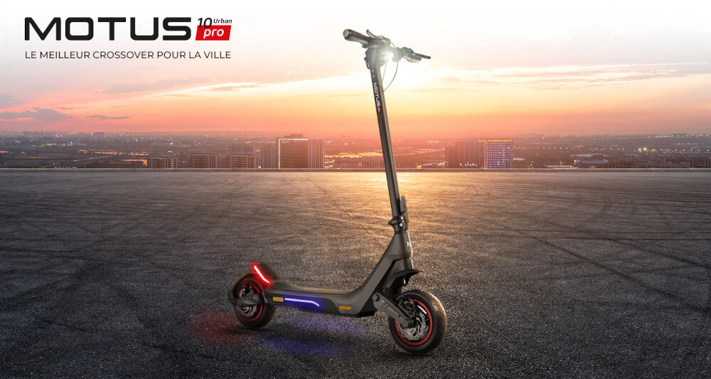 Hulajnoga elektryczna stoi na tle miejskiego krajobrazu o zachodzie słońca. Widoczne jest logo \'MOTUS 10 Urban Pro\' oraz napis \'LE MEILLEUR CROSSOVER POUR LA VILLE\'.