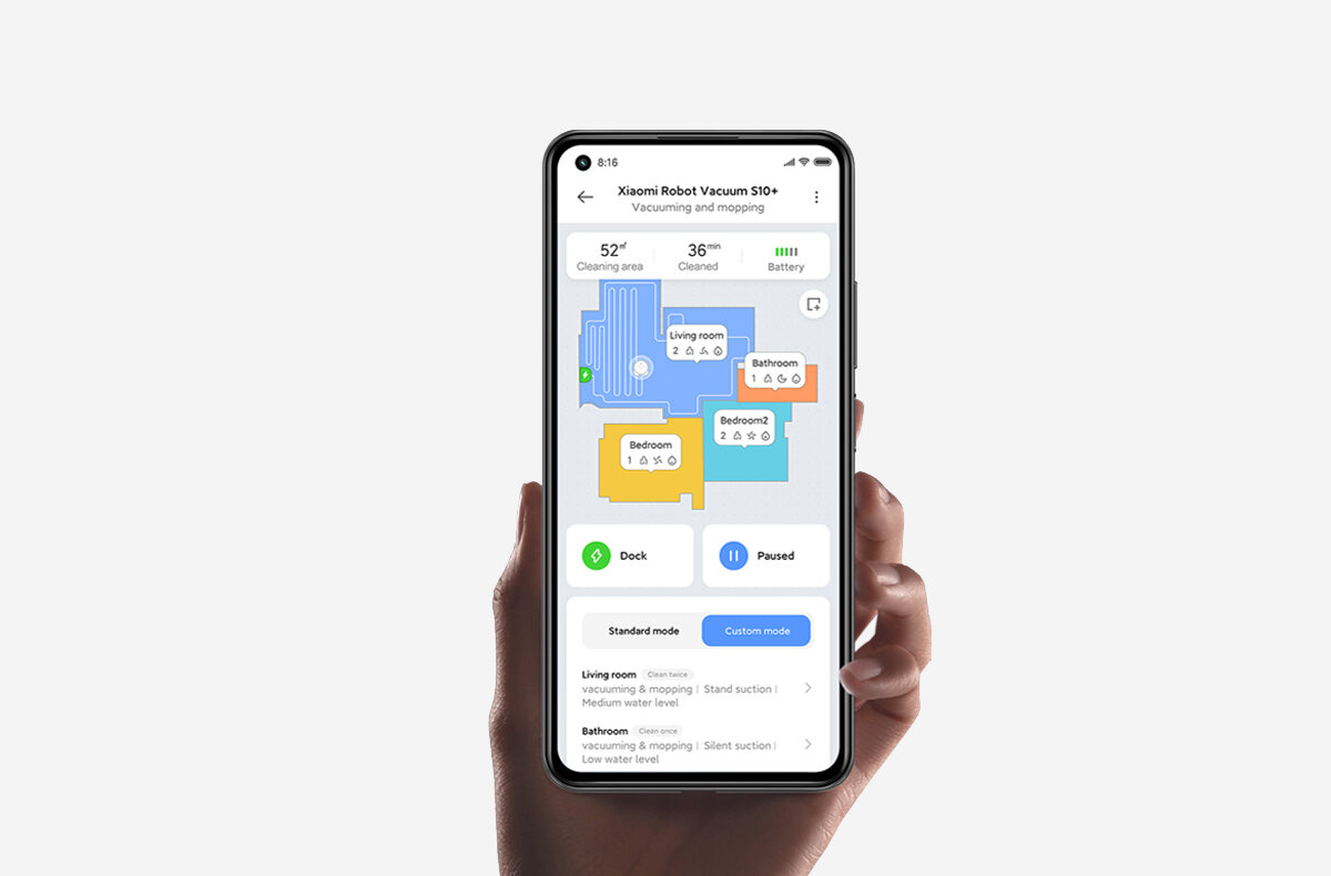 Osoba trzyma smartfon z aplikacją pokazującą mapę sprzątania dla \'Xiaomi Robot Vacuum S10+\'. Na ekranie widoczne są różne pomieszczenia oznaczone kolorami oraz przyciski \'Dock\' i \'Paused\'.