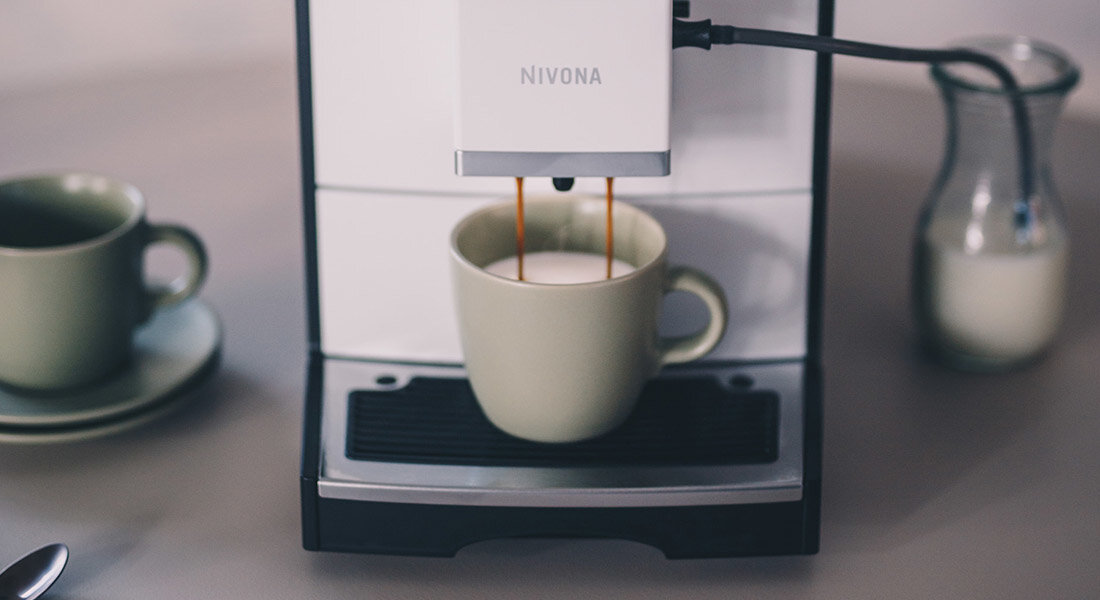 Ekspres do kawy z logo NIVONA nalewa kawę do filiżanki. Obok znajduje się dzbanek z mlekiem.