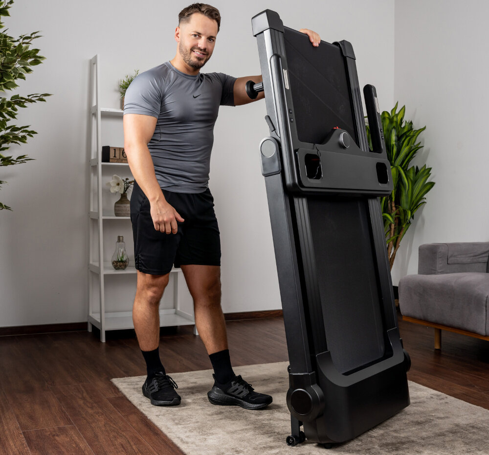 Mężczyzna w sportowym stroju stoi obok złożonej bieżni elektrycznej w jasnym pomieszczeniu. W tle widoczna jest roślina i regał z dekoracjami.