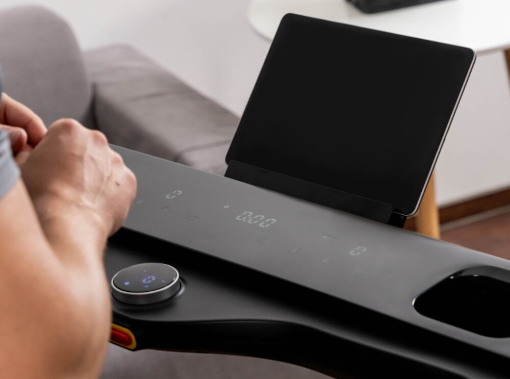 Osoba obsługuje panel sterowania bieżni elektrycznej z wyświetlaczem cyfrowym i pokrętłem. W tle widoczny jest uchwyt na tablet lub telefon.