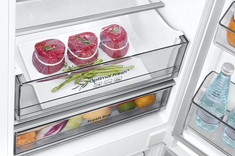 Wnętrze lodówki z szufladą oznaczoną jako \'Optimal Fresh+\', w której znajdują się trzy kawałki mięsa i kilka szparagów. Poniżej widoczna jest szuflada \'Fresh Box\' z owocami i warzywami, a na drzwiach butelki z wodą.