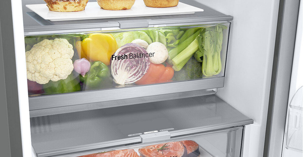 Wnętrze lodówki z widocznymi półkami, na których znajdują się warzywa i owoce w przezroczystych pojemnikach. Na jednym z pojemników widoczny jest napis \'Fresh Balancer\'.