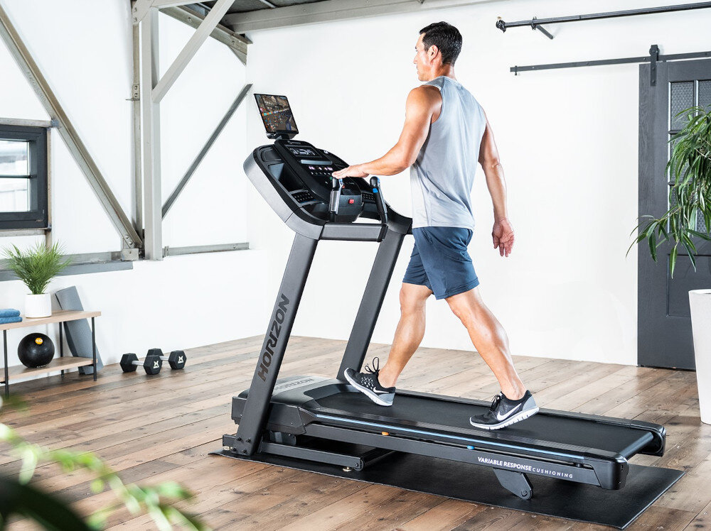 Osoba ćwiczy na bieżni elektrycznej z widocznym logo \'HORIZON\'. W tle znajduje się jasne pomieszczenie z roślinami i sprzętem do ćwiczeń.