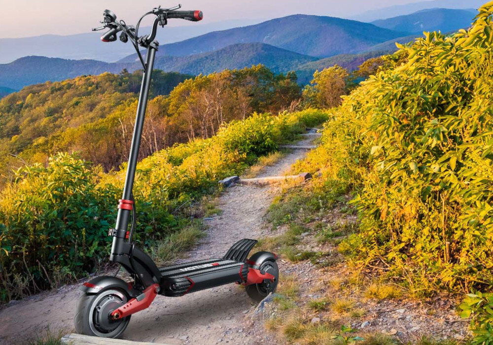 Czarna hulajnoga elektryczna z czerwonymi akcentami stoi na ścieżce wśród zielonych krzewów i górskiego krajobrazu. Hulajnoga ma duże koła i solidną konstrukcję.