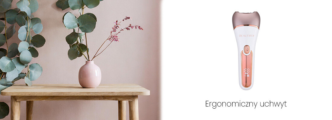 Na obrazie znajduje się depilator w kolorze białym z różowymi elementami oraz napis \'Ergonomiczny uchwyt\'. Obok widoczny jest drewniany stolik z różową wazonem i zielonymi liśćmi.