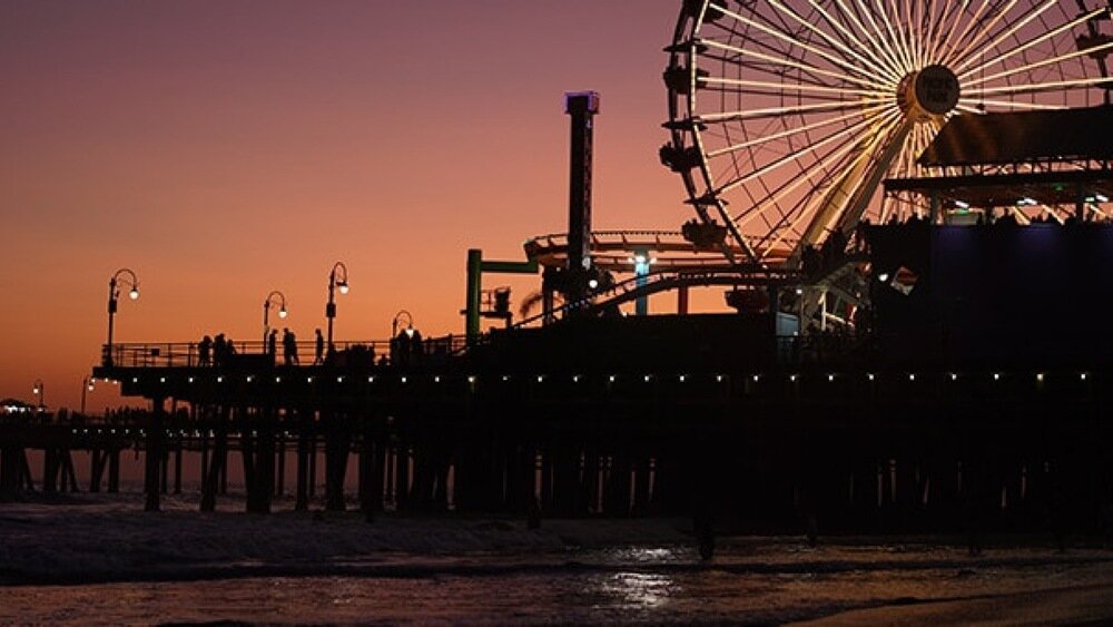Na zdjęciu widać molo z diabelskim młynem oświetlonym światłami na tle zachodzącego słońca. Woda uderza o brzeg, a na molo znajdują się sylwetki ludzi.