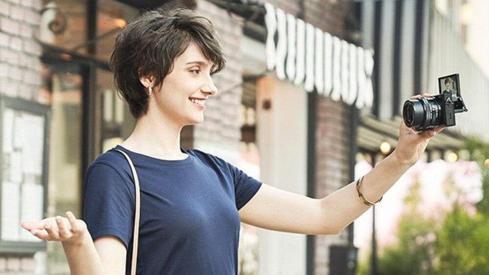 Osoba trzyma aparat bezlusterkowy z odchylanym ekranem, stojąc na zewnątrz przed budynkiem z ceglaną ścianą. W tle widoczne jest rozmyte logo sklepu media expert.