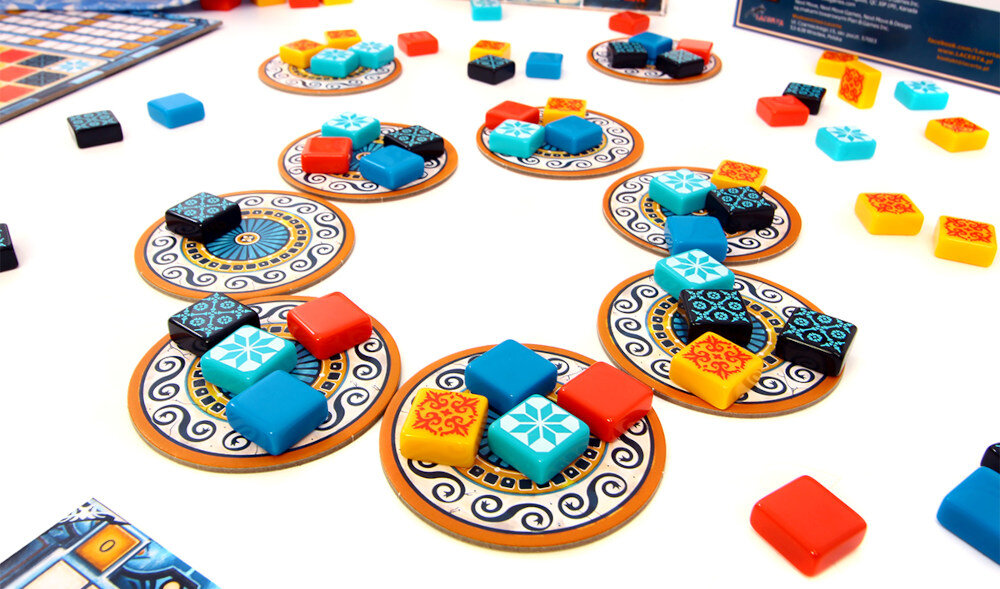 Na obrazie widoczne są elementy gry planszowej z kolorowymi kafelkami w odcieniach niebieskiego, czerwonego, żółtego i czarnego, ułożonymi na ozdobnych okrągłych planszach. W tle znajdują się dodatkowe kafelki i fragmenty planszy gry.