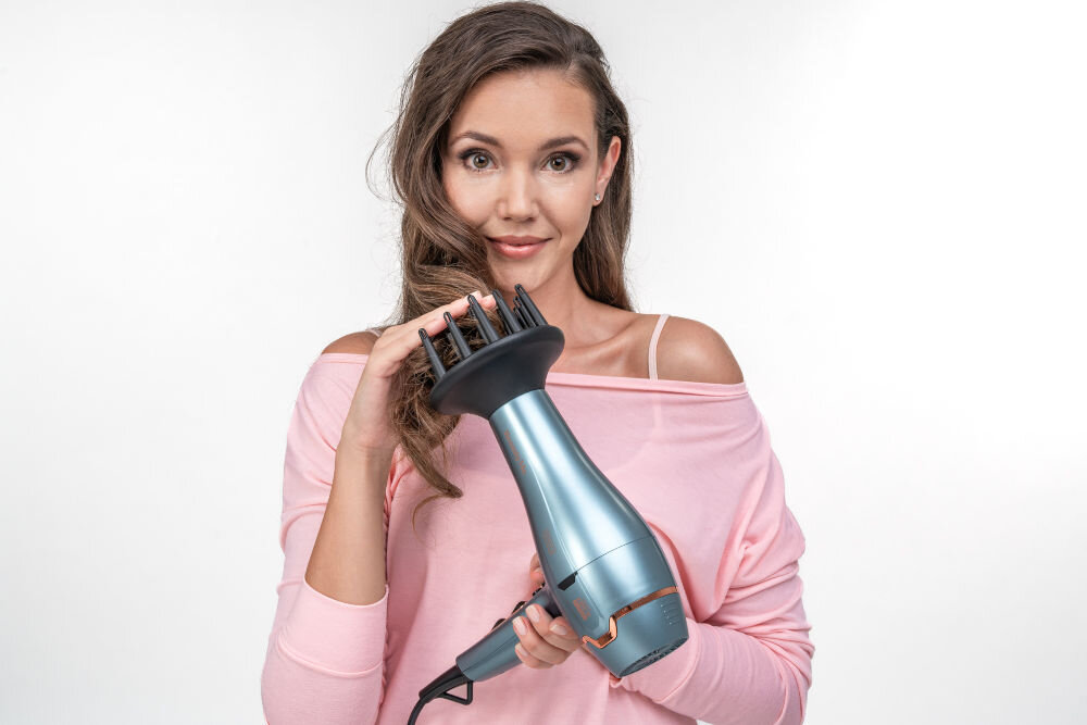 Osoba trzyma niebieską suszarkę do włosów z dyfuzorem. Suszarka ma nowoczesny design i jest skierowana w stronę kamery.