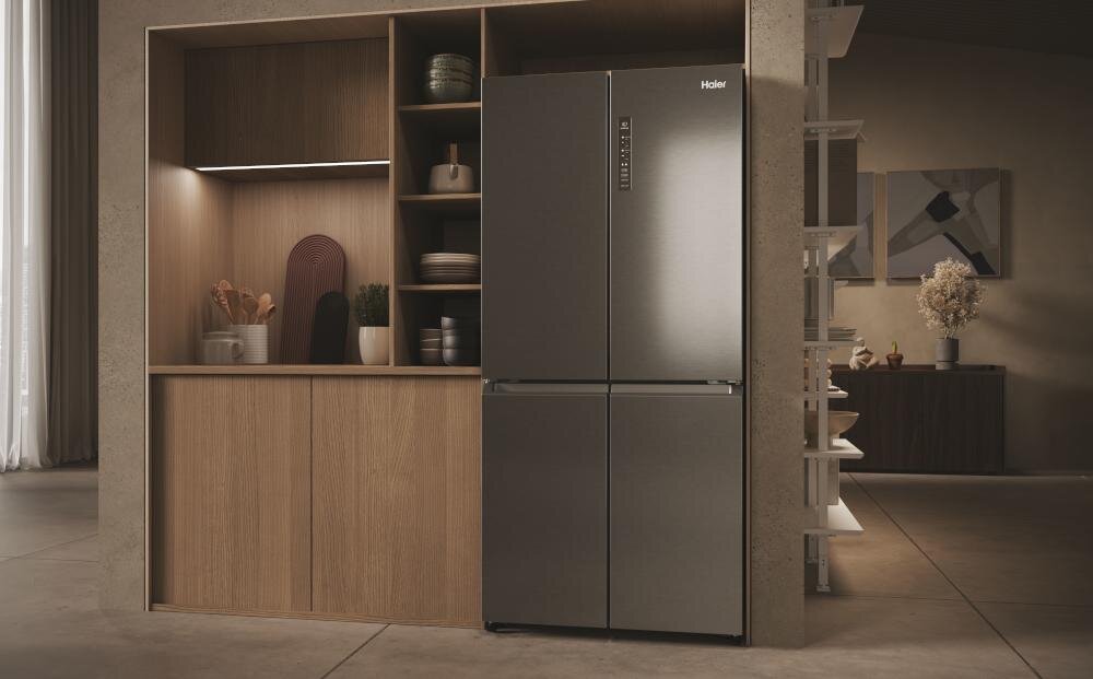 Na obrazie widoczna jest lodówka w kolorze stalowym z logo \'Haier\' na górnych drzwiach. Obok lodówki znajduje się drewniana zabudowa z półkami, na których stoją różne przedmioty kuchenne.