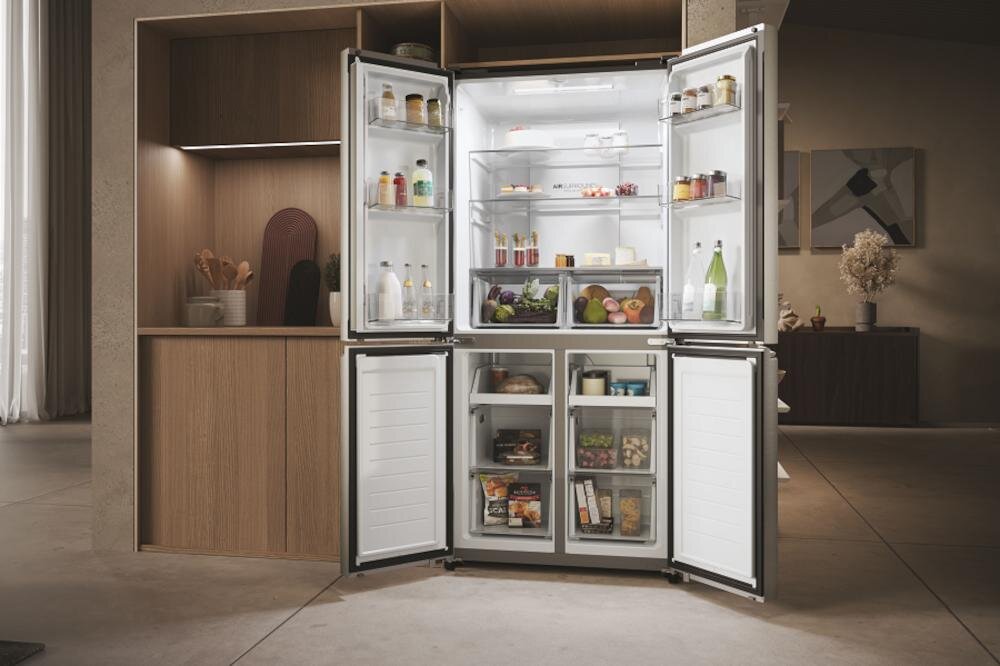 Otwarta lodówka z widocznymi półkami wypełnionymi różnymi produktami spożywczymi, takimi jak napoje, owoce i warzywa. W tle znajduje się nowoczesna kuchnia z drewnianymi szafkami i dekoracjami.