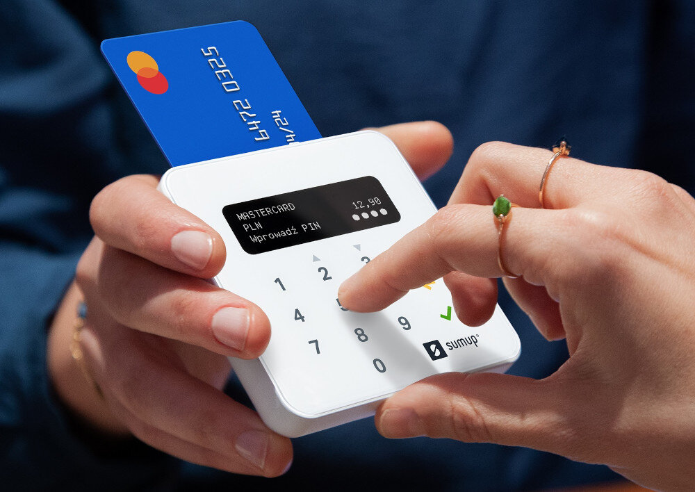 Osoba trzyma terminal płatniczy z kartą włożoną do urządzenia. Na ekranie widoczny jest tekst: \'MASTERCARD PLN 12,90 Wprowadź PIN\'.