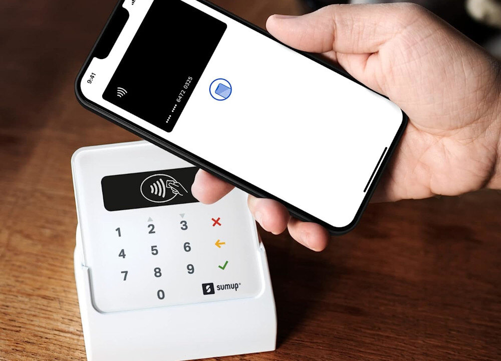 Osoba trzyma smartfon z wyświetlonym ekranem płatności zbliżeniowej nad terminalem płatniczym z logo \'sumup\'. Terminal ma klawiaturę numeryczną i symbole akceptacji oraz odrzucenia transakcji.