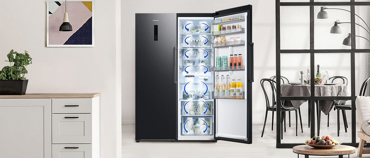 Grafitowa lodówka SAMSUNG z otwartymi drzwiami, ukazująca wnętrze z półkami wypełnionymi napojami i produktami spożywczymi. W tle widoczna jest kuchnia z białymi szafkami i jadalnia z czarnymi krzesłami przy stole.
