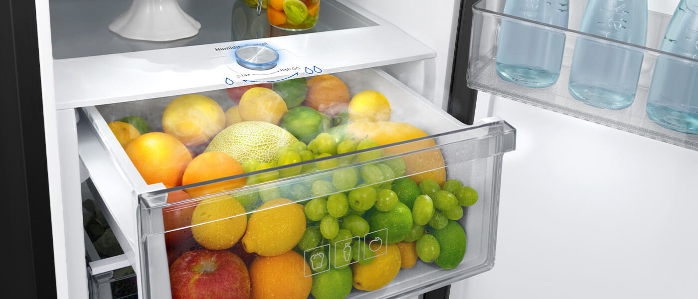 Wnętrze lodówki z przezroczystą szufladą wypełnioną różnymi owocami, takimi jak pomarańcze, winogrona i melon. Na drzwiach lodówki znajdują się butelki z wodą.