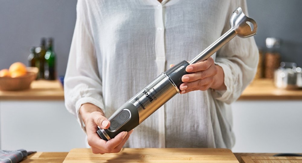 Osoba trzyma metalowy blender ręczny z czarnymi elementami. W tle widać kuchnię z drewnianym blatem i kilkoma przedmiotami.