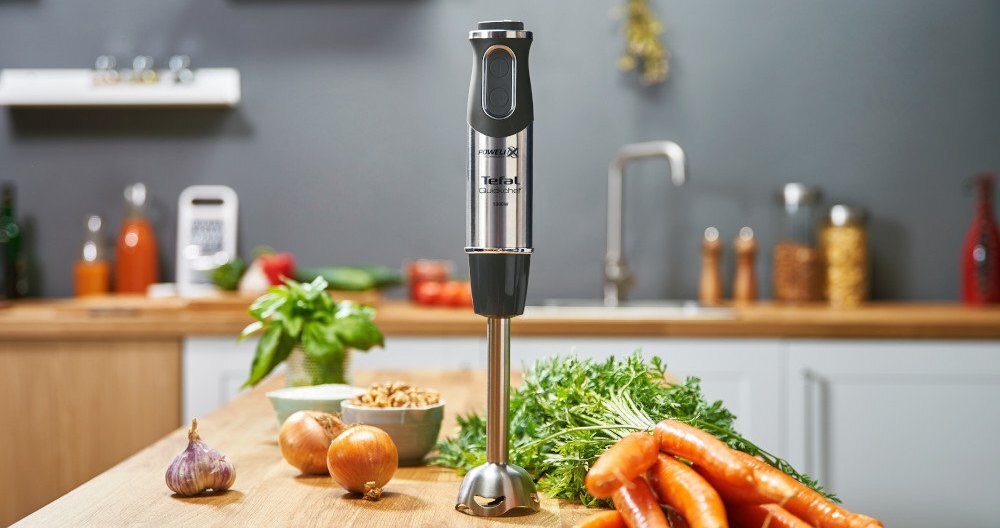 Na blacie kuchennym znajduje się blender ręczny z logo TEFAL, otoczony świeżymi warzywami, takimi jak marchewki i cebula. W tle widoczny jest zlew oraz różne akcesoria kuchenne.