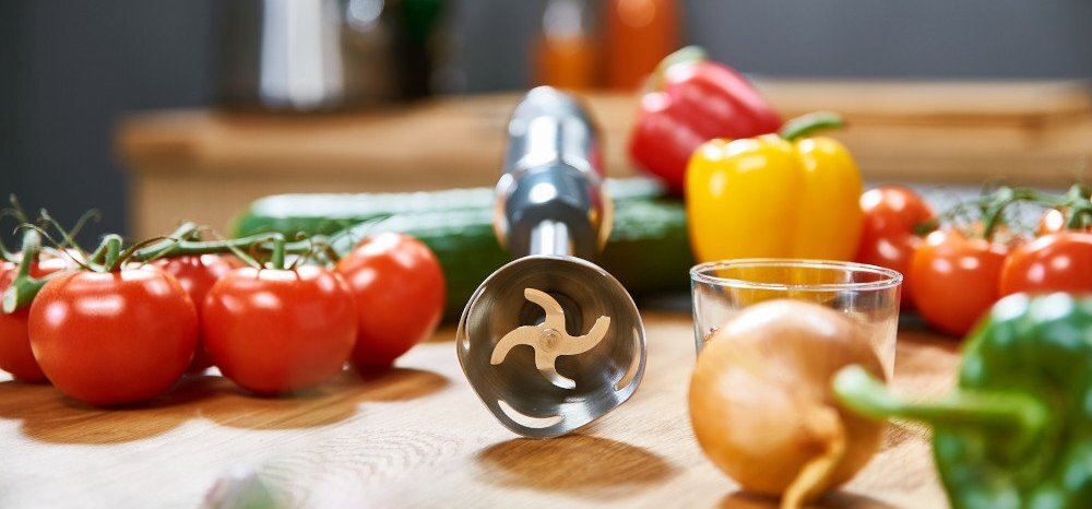 Na drewnianym blacie leży metalowa końcówka blendera z widocznymi ostrzami, otoczona przez różne warzywa, w tym pomidory, papryki i ogórek.