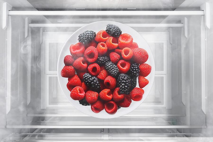 Na obrazie widoczny jest talerz z malinami i jeżynami umieszczony wewnątrz lodówki. Wokół talerza unosi się delikatna mgiełka, sugerująca chłodne wnętrze urządzenia.