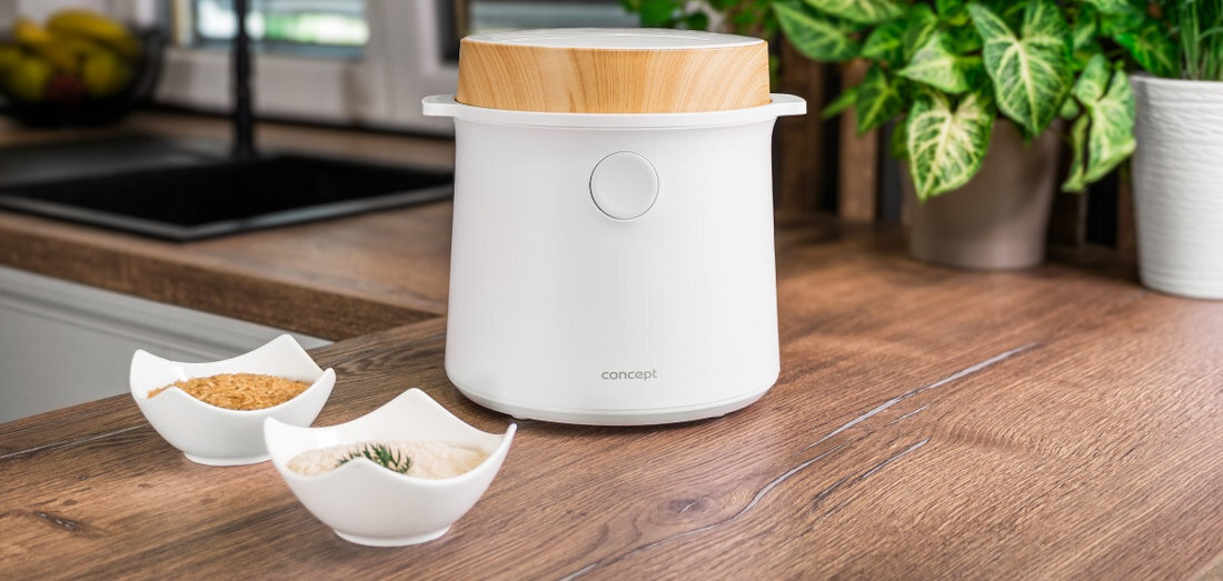 Na drewnianym blacie kuchennym stoi biały ryżowar z drewnianą pokrywką, obok znajdują się dwie miseczki z przyprawami. W tle widoczny jest zlew i roślina doniczkowa.