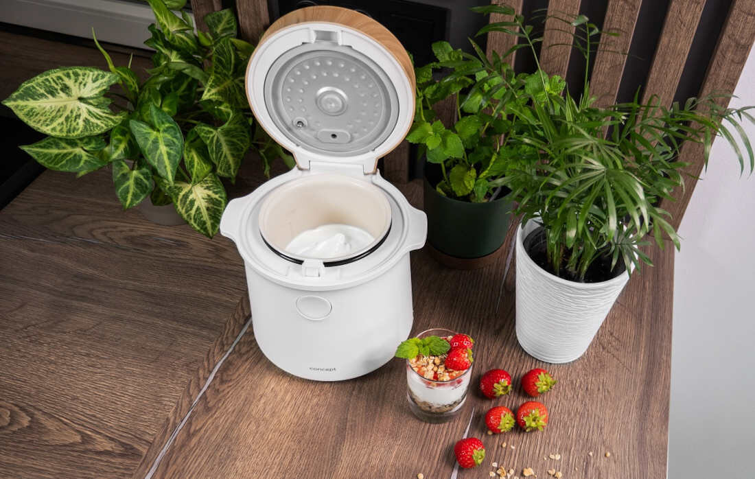 Na drewnianym stole stoi biały ryżowar z otwartą pokrywą, obok którego znajdują się doniczki z roślinami oraz szklanka z deserem i truskawkami. Na urządzeniu widoczne jest logo \'concept\'.