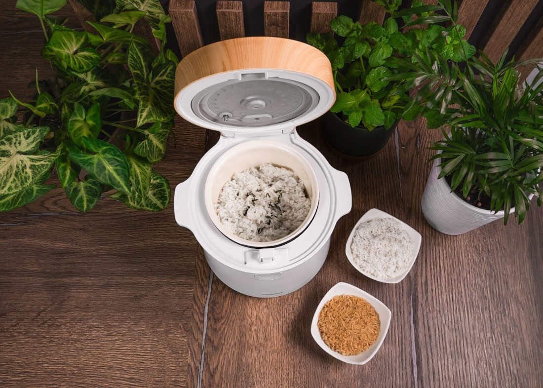 Na drewnianym blacie stoi otwarty ryżowar z ugotowanym ryżem w środku. Obok znajdują się trzy miseczki z różnymi rodzajami ryżu oraz kilka doniczek z zielonymi roślinami.