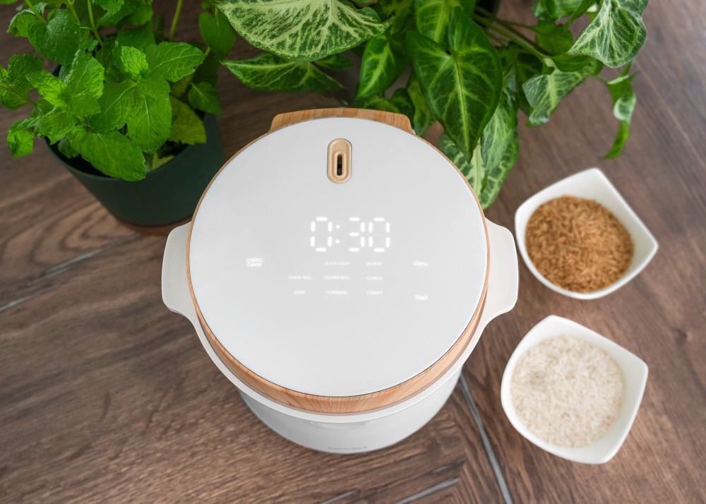 Na drewnianym stole znajduje się biały ryżowar z wyświetlaczem pokazującym \'0:30\'. Obok ryżowara są dwie miseczki z brązowym i białym ryżem oraz doniczka z zieloną rośliną.
