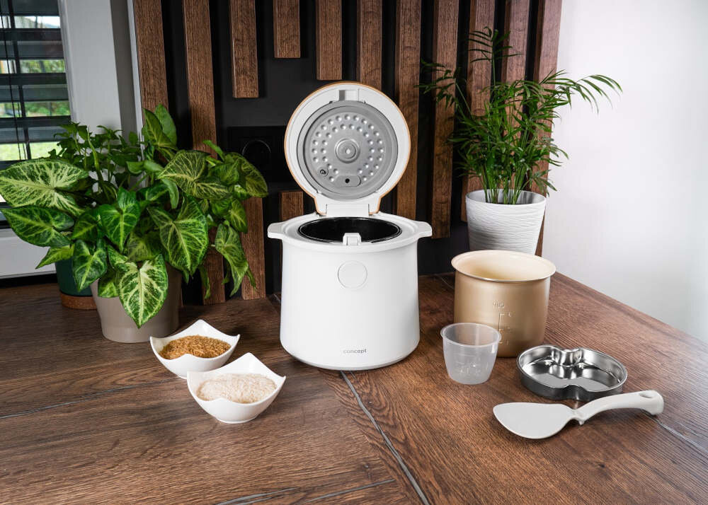 Na drewnianym stole znajduje się biały ryżowar z otwartą pokrywą, obok którego leżą różne akcesoria kuchenne i miseczki z ryżem. W tle widoczne są zielone rośliny doniczkowe oraz drewniana ściana dekoracyjna.