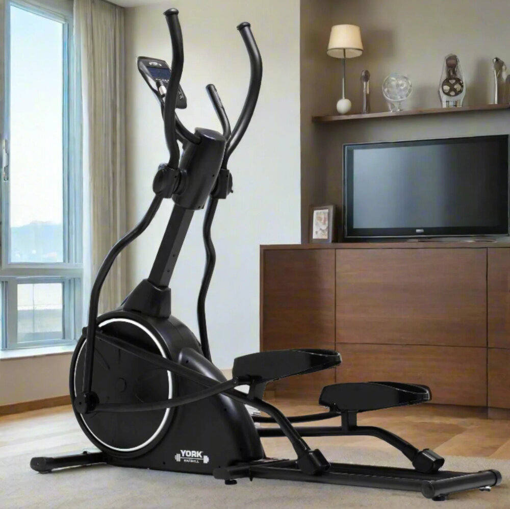 W pokoju znajduje się czarny orbitrek magnetyczny z widocznym logo \'YORK FITNESS\'. W tle widać telewizor na drewnianej szafce oraz półkę z dekoracjami.