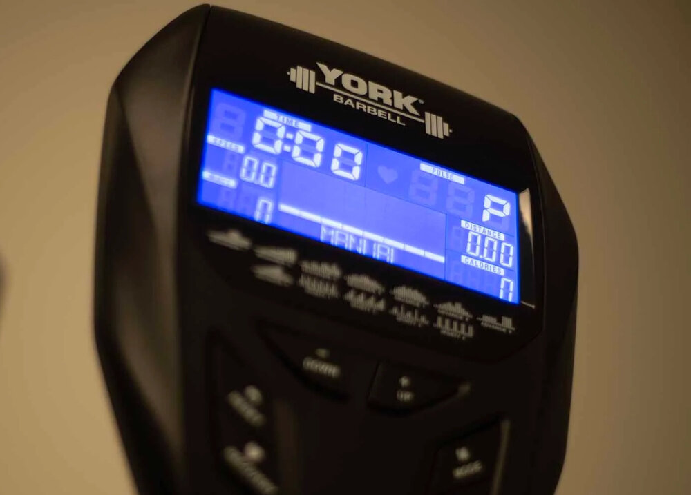 Wyświetlacz urządzenia fitness z logo \'YORK BARBELL\' pokazuje czas 6:50 oraz inne parametry treningu. Ekran jest podświetlony na niebiesko z widocznymi przyciskami poniżej.