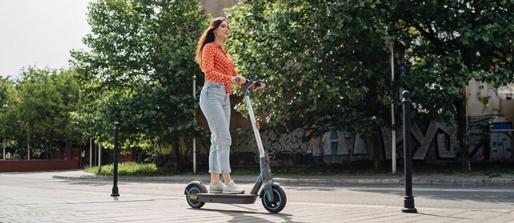 Osoba jedzie na szarej hulajnodze elektrycznej po chodniku w otoczeniu zielonych drzew i budynków. Hulajnoga ma duże koła i nowoczesny design.