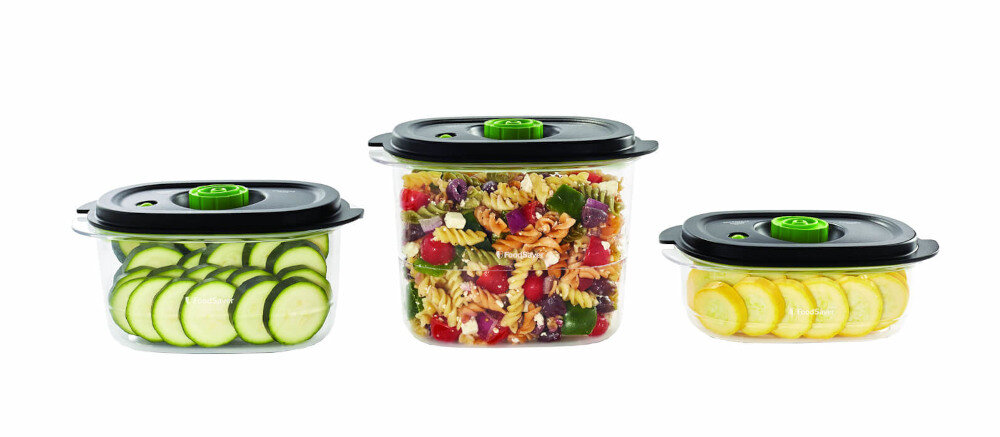 Trzy przezroczyste pojemniki na żywność z czarnymi pokrywkami i zielonymi elementami. W pojemnikach znajdują się pokrojone warzywa i sałatka makaronowa.