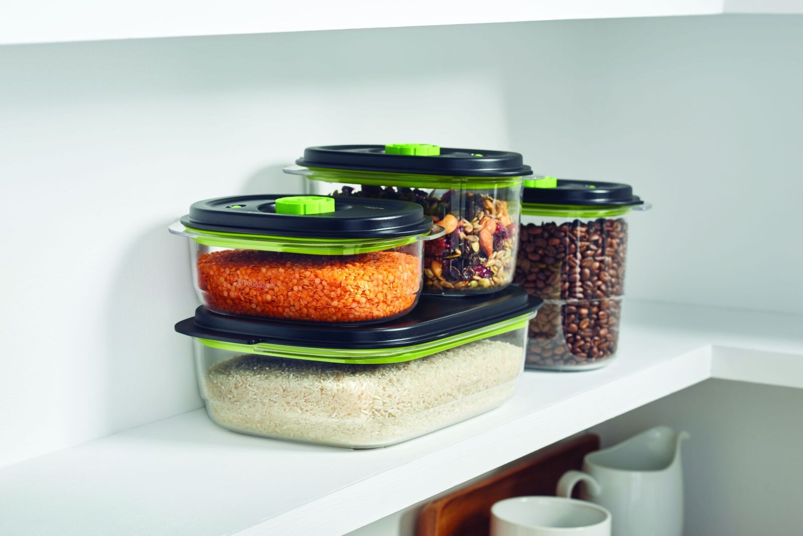 Na półce znajdują się trzy przezroczyste pojemniki na żywność z czarnymi pokrywkami i zielonymi elementami. W pojemnikach widoczne są różne produkty spożywcze, takie jak soczewica, ryż, orzechy i ziarna kawy.