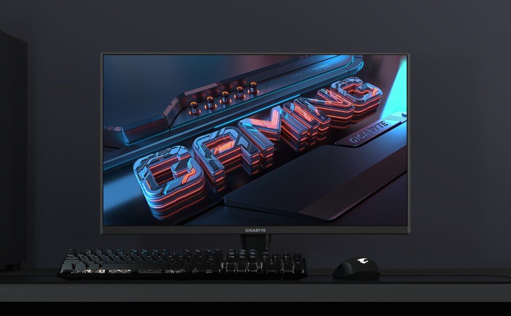 Monitor GIGABYTE GS25F2 Monitor komputerowy ustawiony na biurku obok podświetlanej klawiatury i myszy, wyświetlający futurystyczną grafikę z napisem 'GAMING', opis