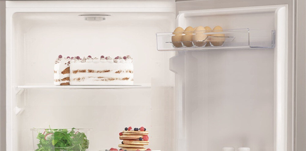 Wnętrze lodówki z widocznymi półkami, na których znajdują się tort, sałatka i naleśniki. Na drzwiach lodówki umieszczony jest pojemnik z jajkami.