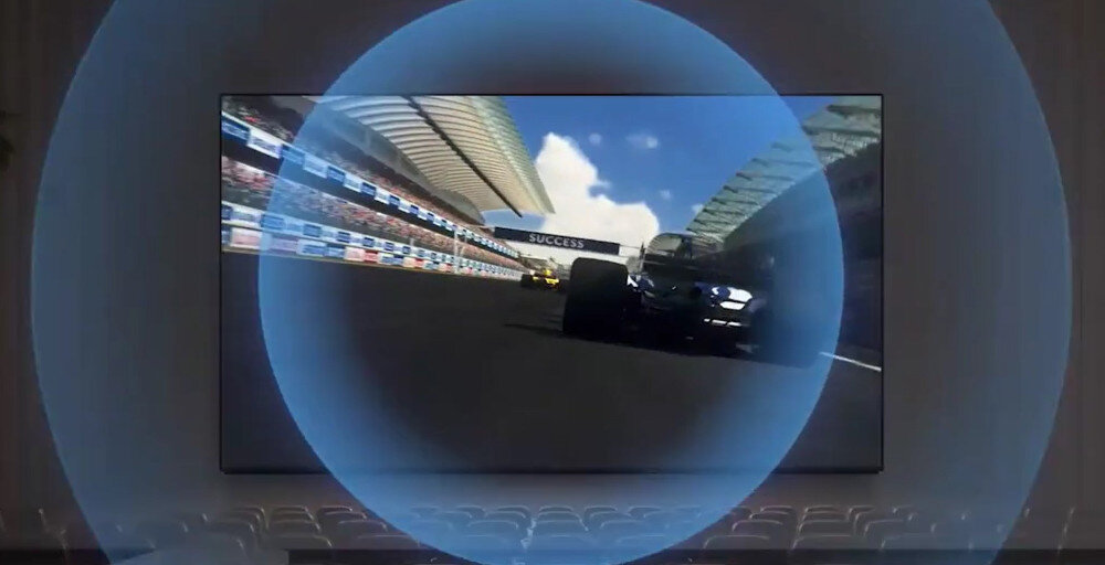Na ekranie telewizora wyświetlana jest scena z wyścigu samochodowego, z widocznym bolidem na torze. W tle widać trybuny i niebo z chmurami.