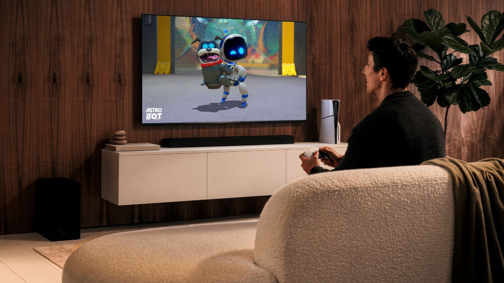 Osoba siedzi na kanapie przed telewizorem, na którym wyświetlana jest gra z postacią robota. Obok telewizora znajduje się konsola do gier, a na ekranie widoczny jest napis \'ASTRO BOT\'.