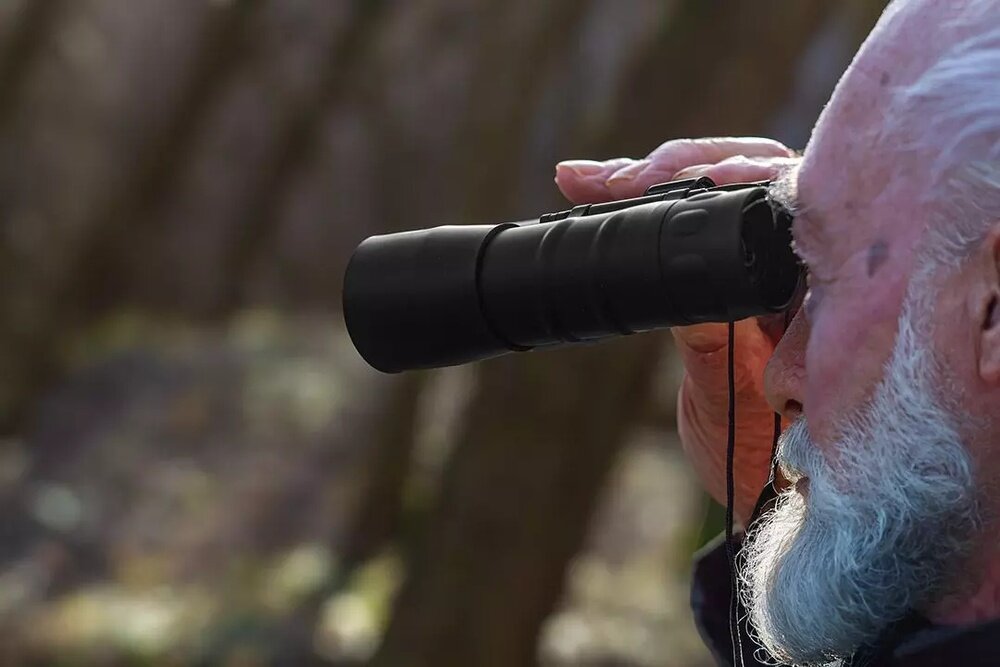 Osoba trzyma lornetkę i patrzy przez nią w dal. W tle widać rozmyty krajobraz z drzewami.