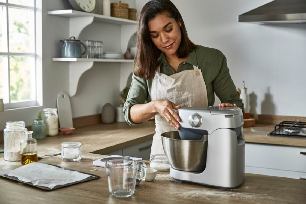 Kobieta w kuchni obsługuje srebrny robot kuchenny planetarny z dużą misą. Na blacie znajdują się różne naczynia i składniki do gotowania.